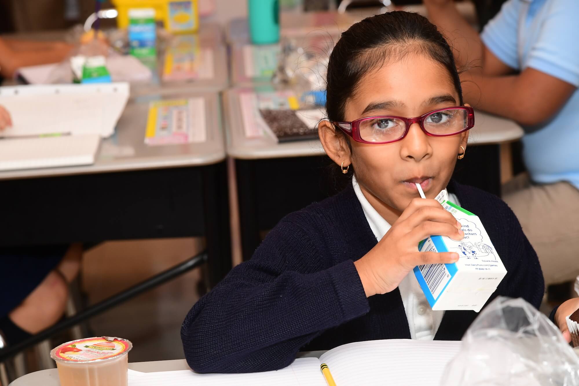 A girl wearing glasses drinks a carton of milk as part of her school lunch.