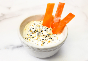 A bowl filled with yogurt dip, seasoning and carrot sticks.