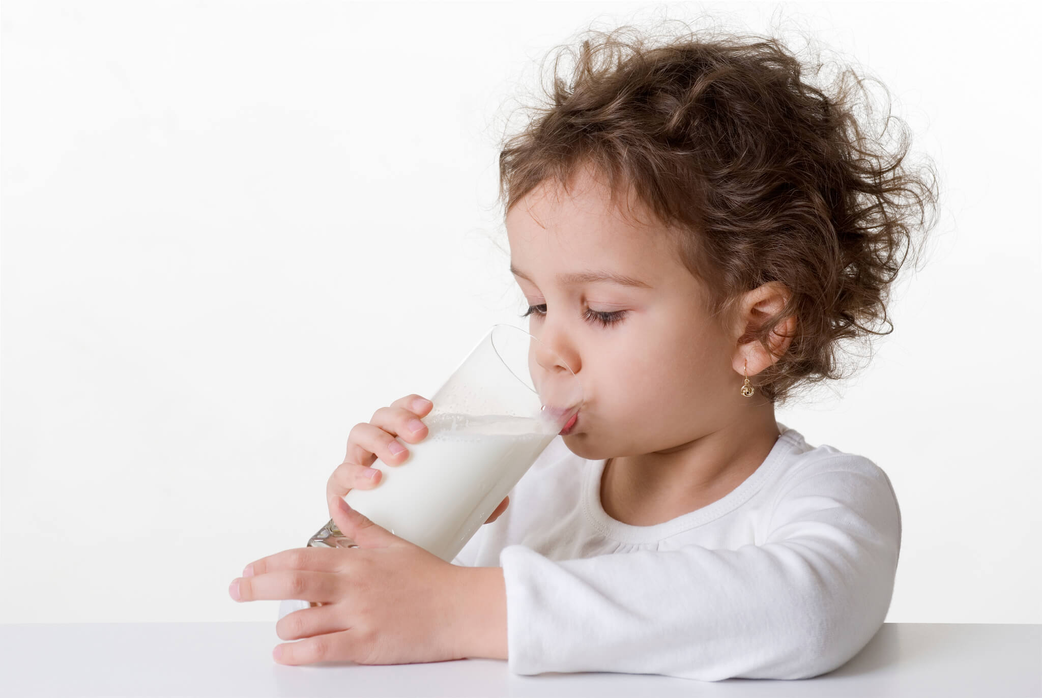 A little girl in a white t-shirt seated on a white table, drinking a glass of milk.