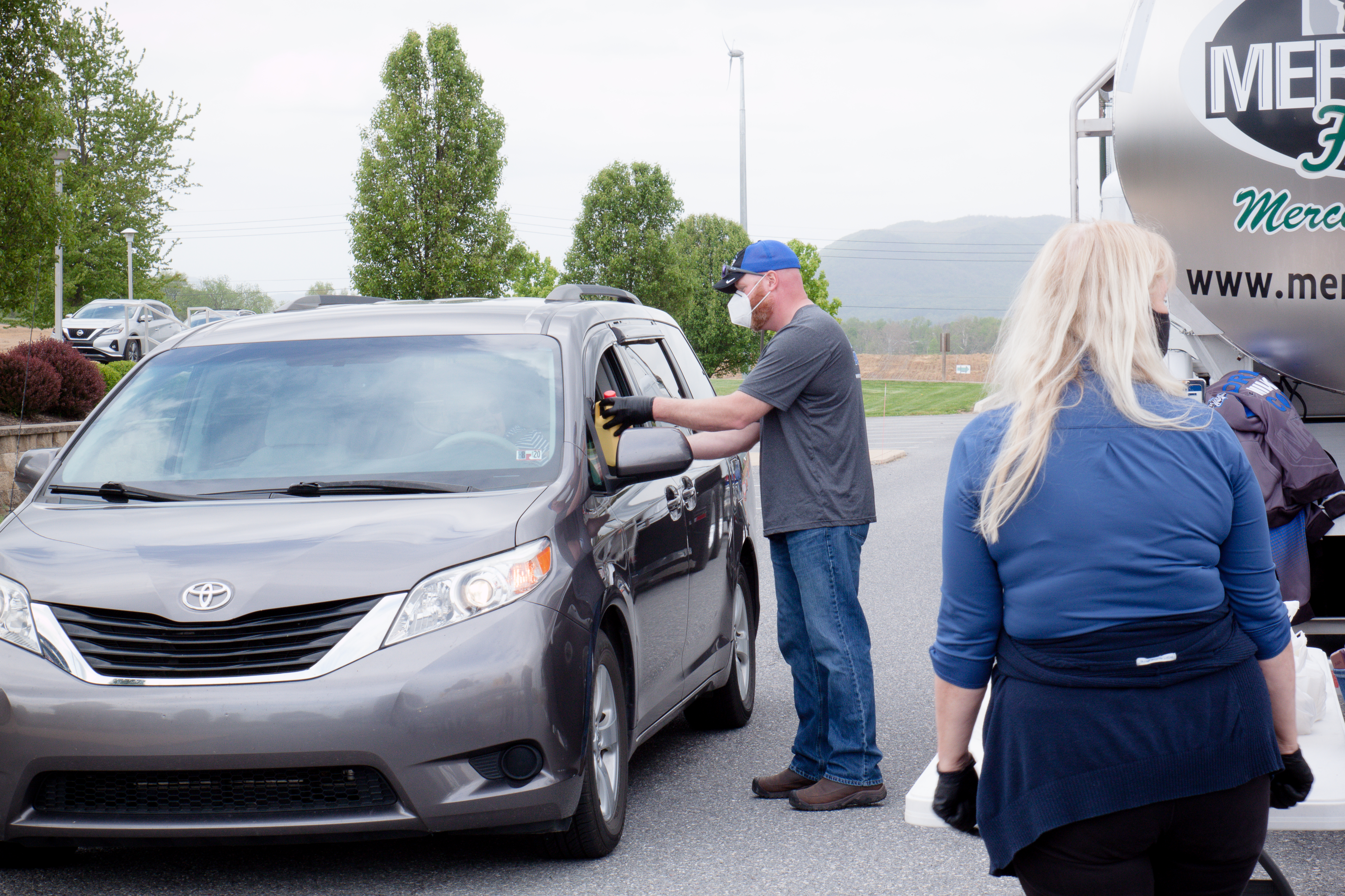 Milk is being delivered to a passenger inside a van by a dairy farmer wearing a mask as part of a food distribution event.