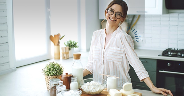 Una mujer frente a una barra de cocina con una botella de leche, un tazón de yogur, un tazón de queso crema, trozos de queso.
