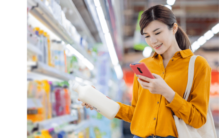 A girl dressed in a yellow shirt shopping in a supermarket and holding a bottle of milk and her cell phone.