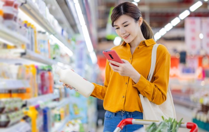 A girl dressed in a yellow shirt shopping in a supermarket and holding a bottle of milk and her cell phone.