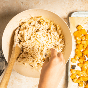 Butternut Squash Pasta Bake Recipe – 3 A woman's hand sprinkling shredded cheese onto a bowl filled with butternut squash pasta.