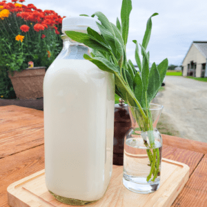 A glass bottle filled with milk placed on a wooden cutting board, accompanied by a vase filled with water and sage.