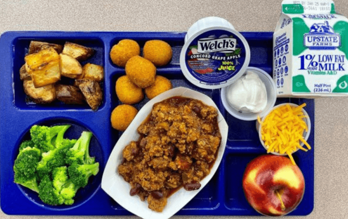 A blue school lunch plate with a carton of milk, grape juice, broccoli, apple, sour cream, cheese, potatoes, and meat.