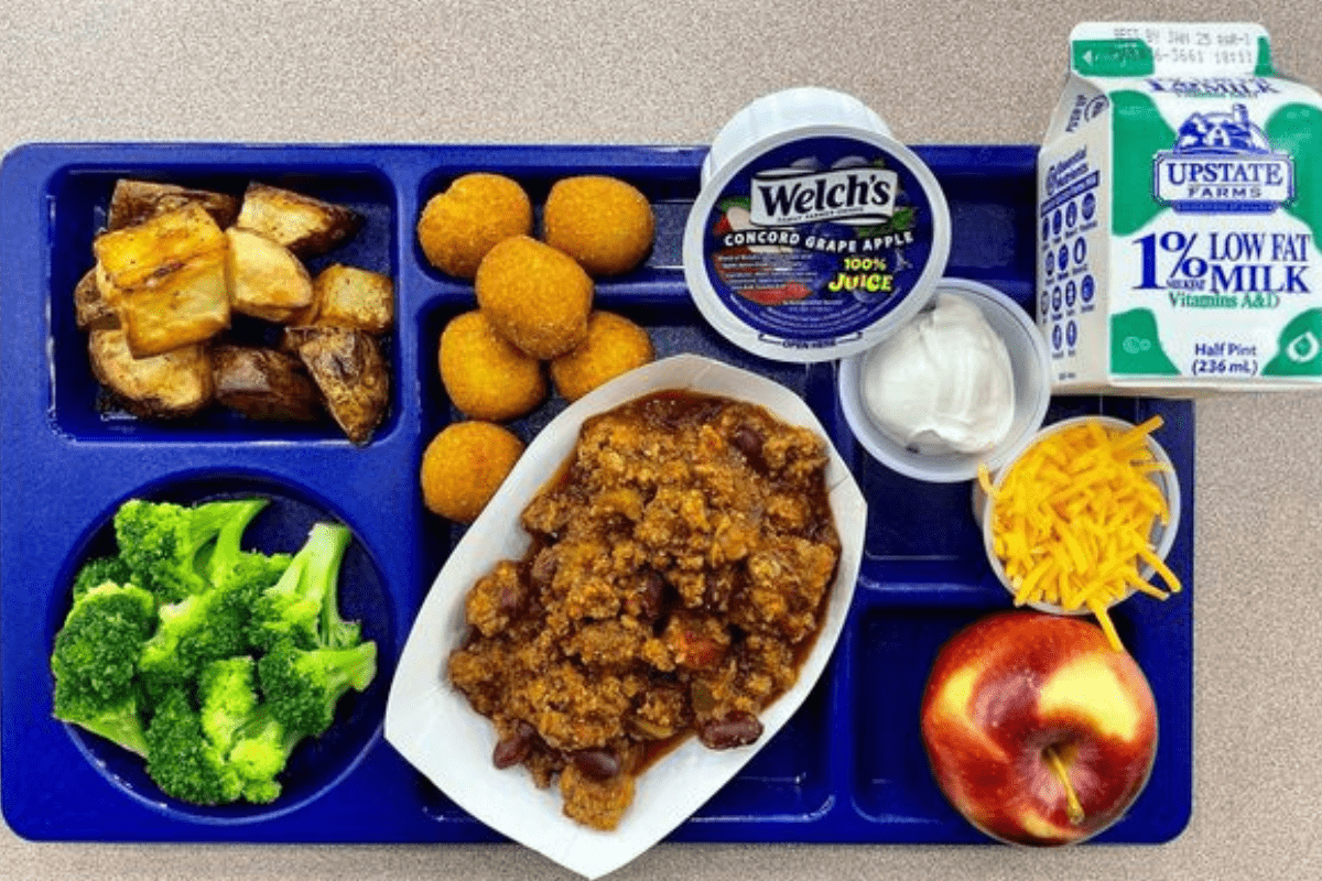 A blue school lunch plate with a carton of milk, grape juice, broccoli, apple, sour cream, cheese, potatoes, and meat.