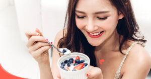 Una mujer sonriente sosteniendo una cuchara a punto de disfrutar de un plato de yogur con arándanos y fresas.
