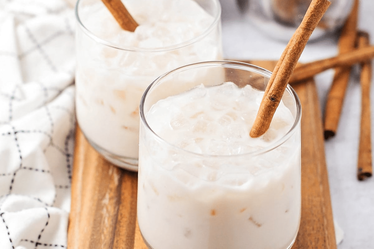 Two glasses with horchata and ice cubes decorated with a cinnamon stick on a wooden board.