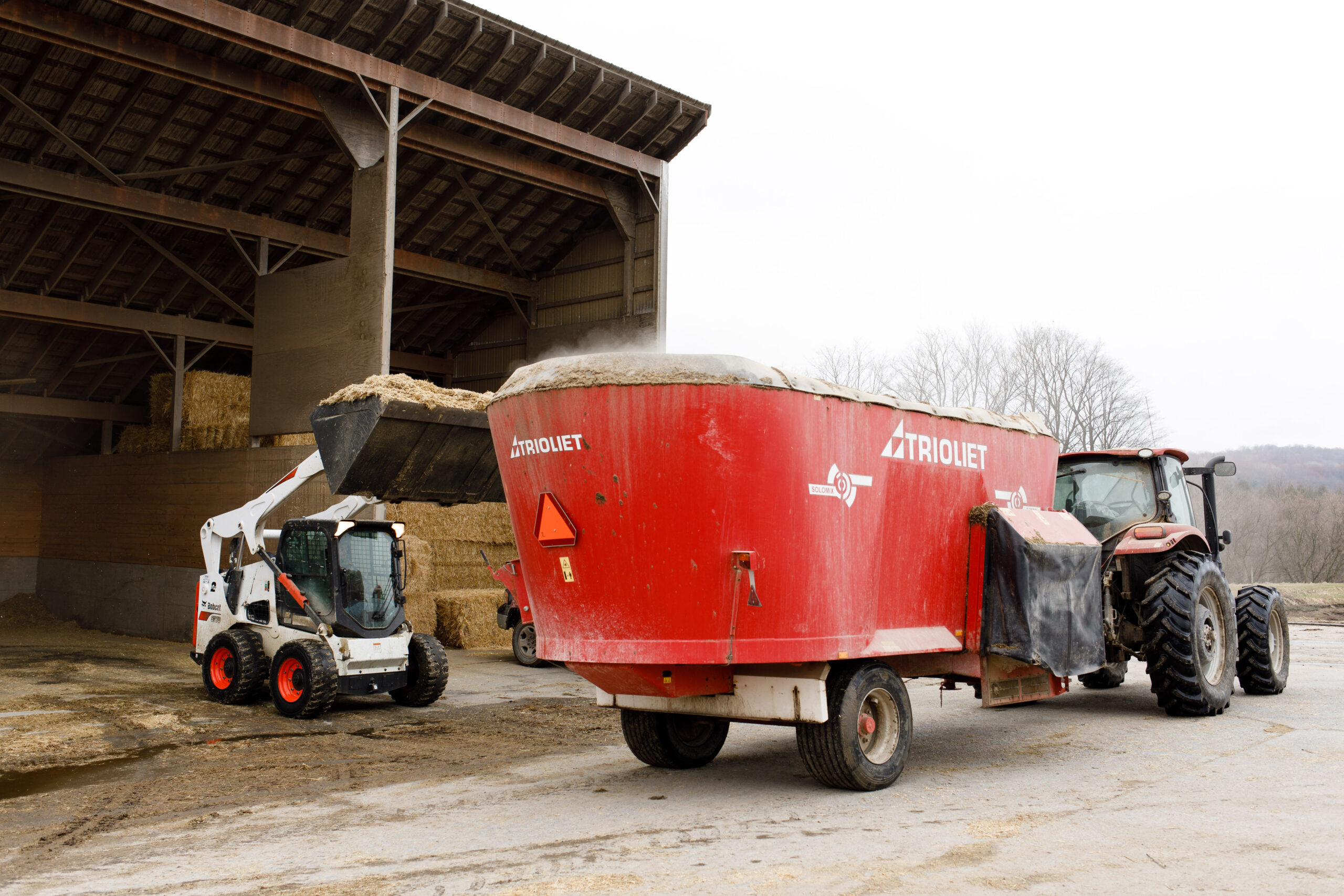 A machine loads TMR feed, for dairy cows, into a red trailer being pulled by a tractor next to a food shed.